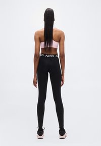Leggings noirs avec une ceinture épaisse arborant un logo blanc, assortis à un brassière de sport rose clair, vus de dos.