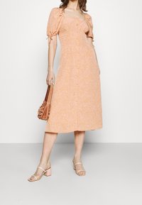 Abito arancione chiaro con scollo a quadrato, maniche a sbuffo e dettagli con bottoni; abbinato a sandali con tacco a blocco rosa e una borsa marrone.
