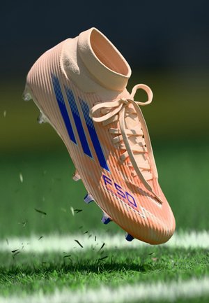 Botas de fútbol de color naranja claro con textura acanalada, rayas azules, cordones blancos y tacos visibles, flotando sobre césped verde.