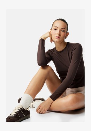 Junge Frau sitzt auf dem Boden, stützt das Kinn auf die Hand, trägt ein braunes Langarmshirt, beige Shorts, weiße Socken und dunkle Sneakers vor weißem Hintergrund.