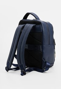 Laivastonsininen reppu, jossa on sileää keinonahkaa ja verkkoviimeistelyjä. Suunniteltu pehmustetuilla säädettävillä olkahihnoilla ja vetoketjullisella osastolla.