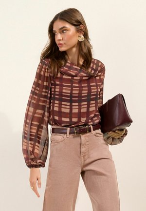 Žena v priesvitnej károvanej blúzke s dlhým rukávom a béžových nohaviciach, drží tmavohnedú koženú listovú kabelku a pozerá sa do strany.