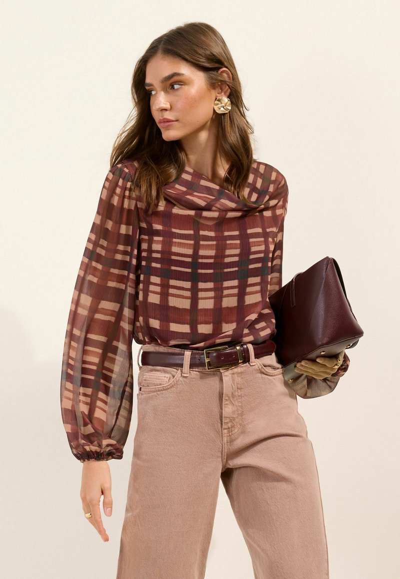 Vrouw draagt een doorschijnende blouse met lange mouwen en ruitpatroon, beige broek, houdt een donkerbruine leren clutch vast en kijkt opzij.