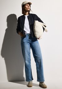 Veste de bombardier marine, t-shirt blanc, jeans droits bleu clair, baskets beiges et sac matelassé crème, posés sur un fond neutre.