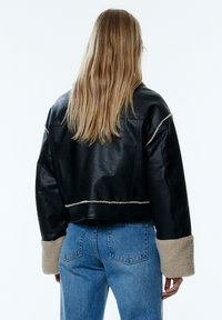 Schwarze Leder-Bomberjacke mit beigem Schafswollbesatz an den Manschetten und dem Saum, mit einfachem Design und lässiger Silhouette. Blaue Jeans sind sichtbar.