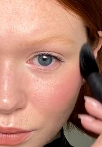 Gros plan sur un visage au teint clair, avec des taches de rousseur légères, des yeux bleus et des joues roses. Un pinceau de maquillage noir est appliqué près de la joue.