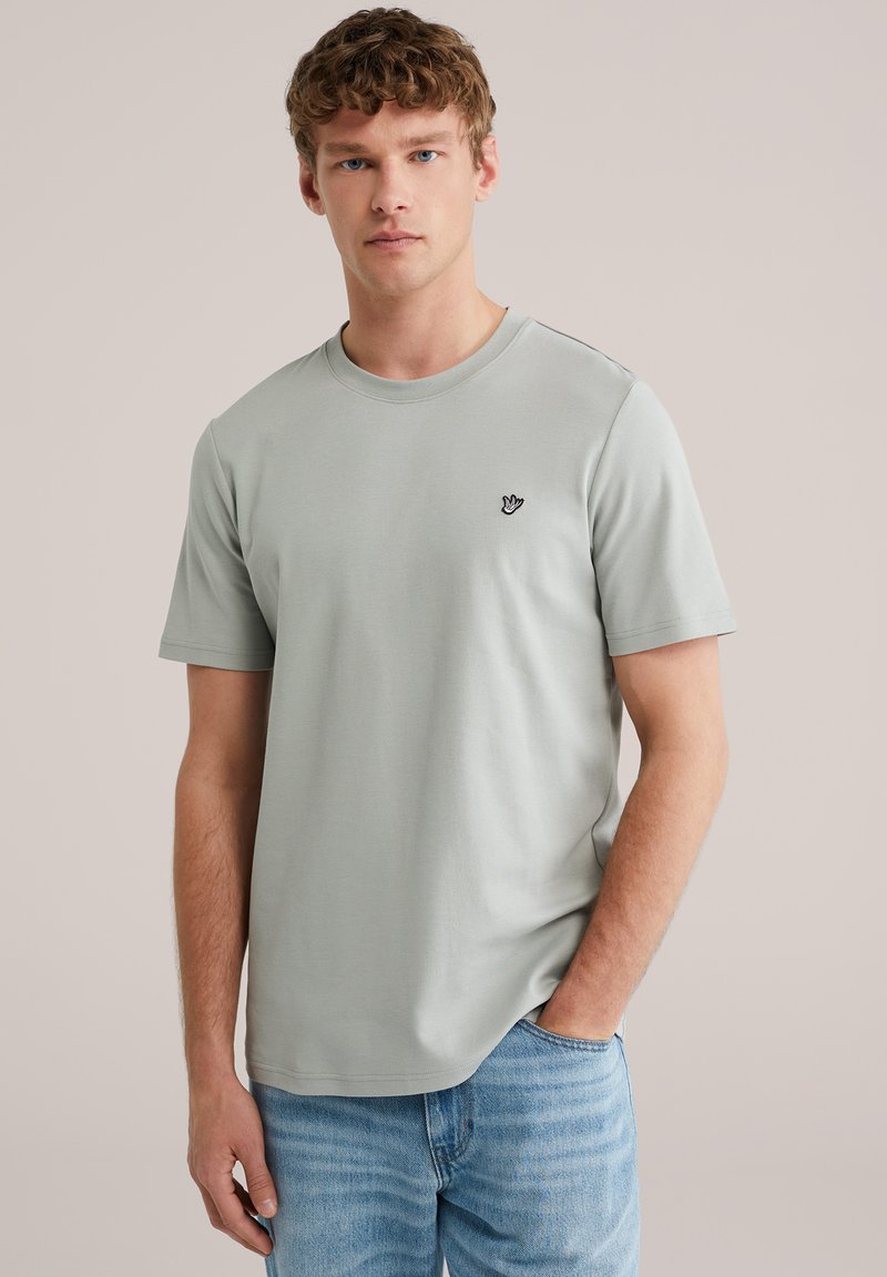 Jonge man met lichtbruin krullend haar, gekleed in een lichtgrijs T-shirt met korte mouwen en een blauwe spijkerbroek, staand met één hand in zijn zak.