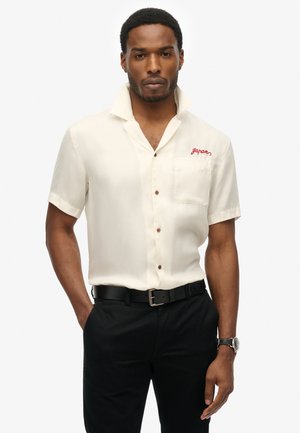 Camicia a maniche corte color crema con colletto a camicia, bottoni marroni, taschino sul petto sinistro e testo ricamato rosso "Giappone." Abbinata a pantaloni neri e cintura.