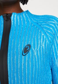 Stickad jacka i ljusblått med silverränder. Har en svart dragkedja och logotypdetalj på framsidan. Texturerad tyg ger ett unikt utseende.