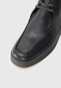 Gros plan sur une chaussure en cuir noir texturé avec des lacets, à bout carré et semelle en caoutchouc épaisse sur fond blanc.