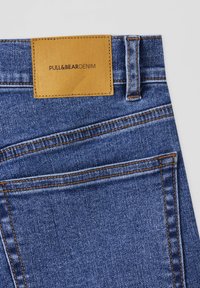 Zone de la poche arrière d'un jean en denim bleu avec des coutures orange et une étiquette en cuir beige portant l'inscription "PULL&BEAR DENIM".