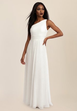 Femme aux longs cheveux raides portant une robe blanche asymétrique longue jusqu'au sol, debout avec une main sur la hanche devant un fond beige.