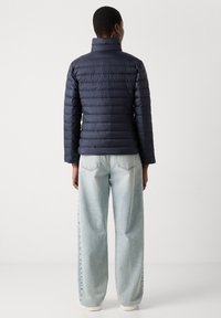 Navy puffert jakke med vandret quiltet design, høj krave og letvægtsmateriale, kombineret med lyseblå jeans med vide ben.