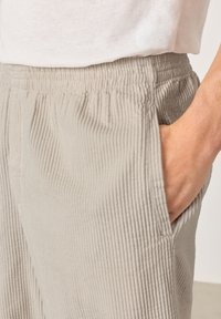 Ręka w kieszeni beżowych spodni sztruksowych z elastycznym pasem, noszonych z białą koszulą.