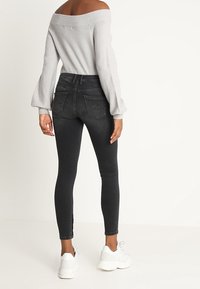 Grauer schulterfreier Pullover und schwarze Skinny-Jeans mit seitlichen Schlitzen, kombiniert mit weißen Sneakers. Die Jeans haben Gesäßtaschen und eine dezente Ausbleichung.