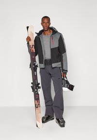 Conjunto de esquí que incluye una chaqueta negra y gris con detalles rojos, una parte superior gris, pantalones oscuros y botas de esquí negras, sosteniendo esquís y un casco.