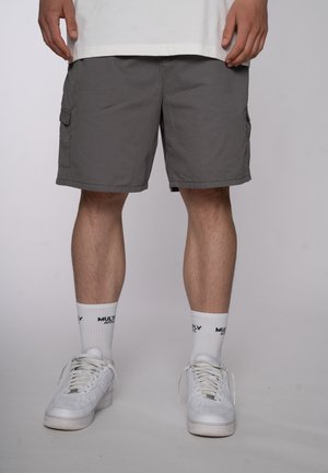Hombre que lleva pantalones cortos cargo grises, calcetines blancos con texto negro y zapatillas blancas, de pie contra un fondo claro y liso.
