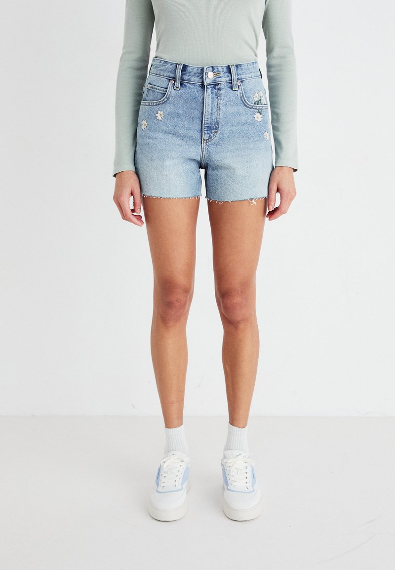 Shorts en denim bleu clair avec un ourlet effiloché, ornés de fleurs brodées sur les poches. Associés à des baskets blanches et un haut à manches longues vert clair.