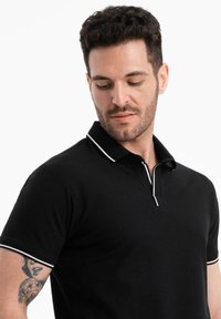 Polo shirt noir en tissu texturé avec un col et des accents de manches à rayures blanches. Équipé d'une patte de boutonnage à trois boutons au niveau du cou.