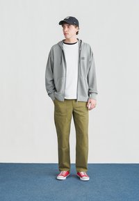 Νέος άνδρας που φοράει γκρι φούτερ με φερμουάρ, λευκή μπλούζα, χακί παντελόνι, κόκκινα αθλητικά παπούτσια και μαύρο ριγέ καπέλο μπέιζμπολ, στέκεται πάνω σε μπλε χαλί.