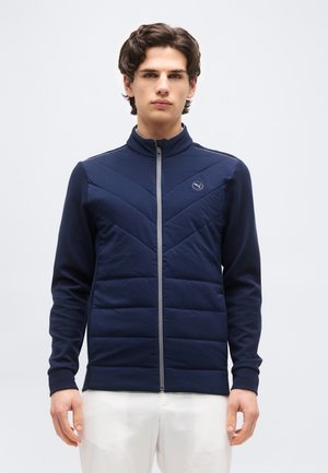 Fiatal férfi, aki sötétkék, steppelt cipzáras dzsekit visel kis Puma logóval és fehér nadrágot, egy sima, világos háttér előtt áll.