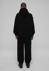 Un sweat à capuche noir avec une grande capuche et un ourlet côtelé, associé à un pantalon de survêtement noir ample. Le matériau semble doux et texturé. Des baskets noires.