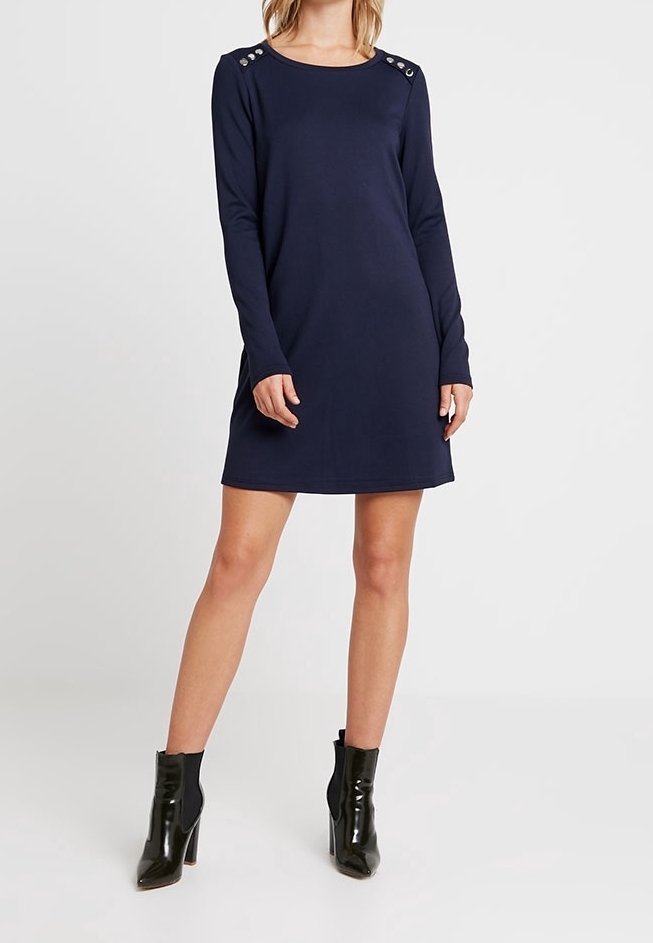 Femme vêtue d'une robe bleu marine à manches longues avec des détails de boutons sur les épaules et des bottines noires à talons hauts, debout sur un fond blanc.