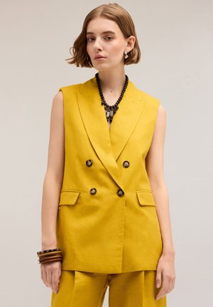 Dubbelblijvende mouwloze blazer in mosterdgele stof, met twee voorzakken, zwarte knopen en een getailleerde pasvorm. Accentuatie met een zwarte kralenketting.