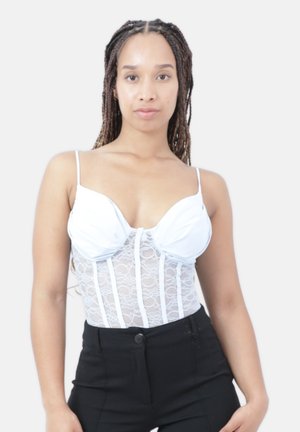 Vrouw met lang gevlochten haar draagt een witte kanten bustiertop met dunne bandjes en een zwarte high-waisted broek, staand tegen een effen achtergrond.