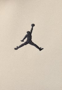 Logo Jumpman brodé en noir sur un tissu beige texturé, représentant une figure dans une pose dynamique tenant un ballon de basket.