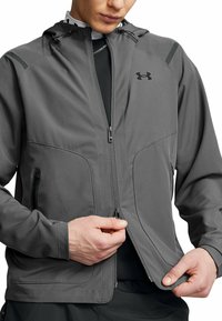Veste grise zippée avec capuche, présentant une texture lisse, des poches zippées et le logo Under Armour sur la poitrine. Cordon de serrage réglable sur la capuche.