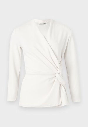 Λευκό μπλούζα με μακριά μανίκια, τύπου wrap, με V-λαιμόκοψη και λεπτομέρεια κόμπου στο πλάι, σε εφαρμοστή γραμμή, εμφανιζόμενη σε απλό ανοιχτό γκρι φόντο.