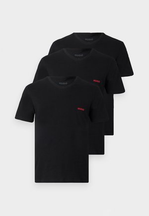 Homme debout pieds nus, portant un t-shirt noir à manches courtes avec un petit logo rouge et un short bleu texturé sur fond blanc uni.