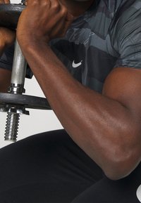 Camiseta deportiva negra con patrón de camuflaje gris, sosteniendo una barra metálica, mostrando un brazo musculoso con textura definida y brillo.