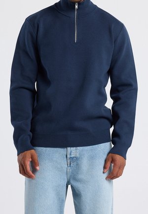 Mand iført marineblå sweater med kvartlyn-lukning og lyseblå jeans, stående med hænderne afslappet ned langs siderne.