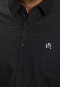 Chemise noire à boutons avec un petit motif blanc et une poche poitrine ornée d'un logo brodé blanc avec des lignes croisées et des lettres.