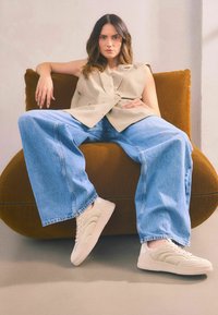 Veste sans manches beige, jean large bleu clair, et baskets crème avec des détails texturés, assis sur une chaise arrondie en velours côtelé marron.