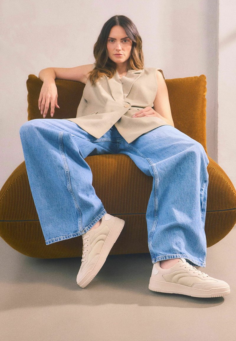 Beige ärmelloser Blazer, hellblaue weit geschnittene Jeans und cremefarbene Sneakers mit strukturierten Details, sitzen auf einem runden braunen Cordstuhl.