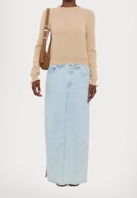 Femme portant un pull beige à manches longues, une jupe longue en denim bleu clair avec une couture frontale, et portant un sac à bandoulière marron.
