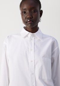 White cotton button-up shirt with a collar, long sleeves, a patch pocket on the left, and a smooth texture.