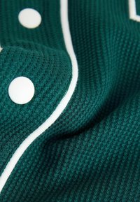 Textúrovaná tmavozelená látka so vzorom mriežky, doplnená o biele lemovanie a veľké biele gombíky. Detailný záber zobrazujúci záhyby a detaily.