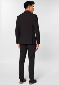 Abito nero con vestibilità su misura, caratterizzato da una fessura singola e rever a lancia. Camicia rossa sotto, abbinata a scarpe di pelle nere.