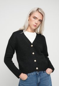 Juodas megztinis su trumpu kirpimu, turintis V formos iškirpimą ir penkias dideles kreminės spalvos mygtukus, derinamas su baltu marškinėliu ir mėlynomis džinsų kelnėmis.