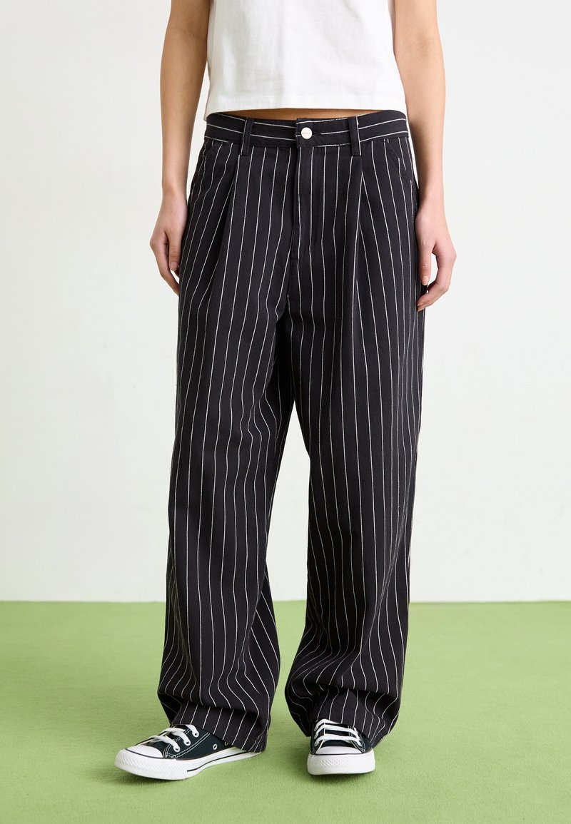 Personne portant un pantalon noir large à fines rayures et des baskets basses noires, debout sur un sol vert face à un mur blanc.