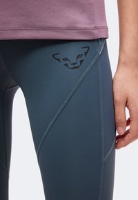 Gros plan d'une personne portant des leggings bleu foncé avec un logo noir stylisé représentant un visage d'animal sur la cuisse et un haut mauve.