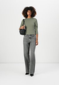 Olijf groene geribbelde lange mouwen top gecombineerd met hoge taille, losse grijze jeans en zwarte puntige schoenen, terwijl ze een kleine zwarte handtas vasthoudt.