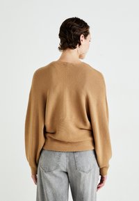 Kvinne med kort hår som har på seg store hoopsmykker, en løs beige ribbestrikket genser og lysegrå høy midje jeans, vendt bort fra hvit bakgrunn.