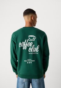 Groene sweatshirt met witte tekst en graphics op de achterkant: "koffieclub" en een illustratie van een koffiekop. Casual fit, zachte stof, ronde hals.