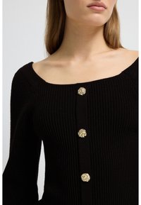 Svart ribbstickad cardigan med fyrkantig urringning och tre dekorativa guldfärgade knappar framtill. Mjuk textur med subtil glans.