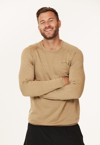 Beige langærmet skjorte med tekstureret overflade og rund halsudskæring, der har et diskret logo på brystet. Enkel, atletisk pasform.